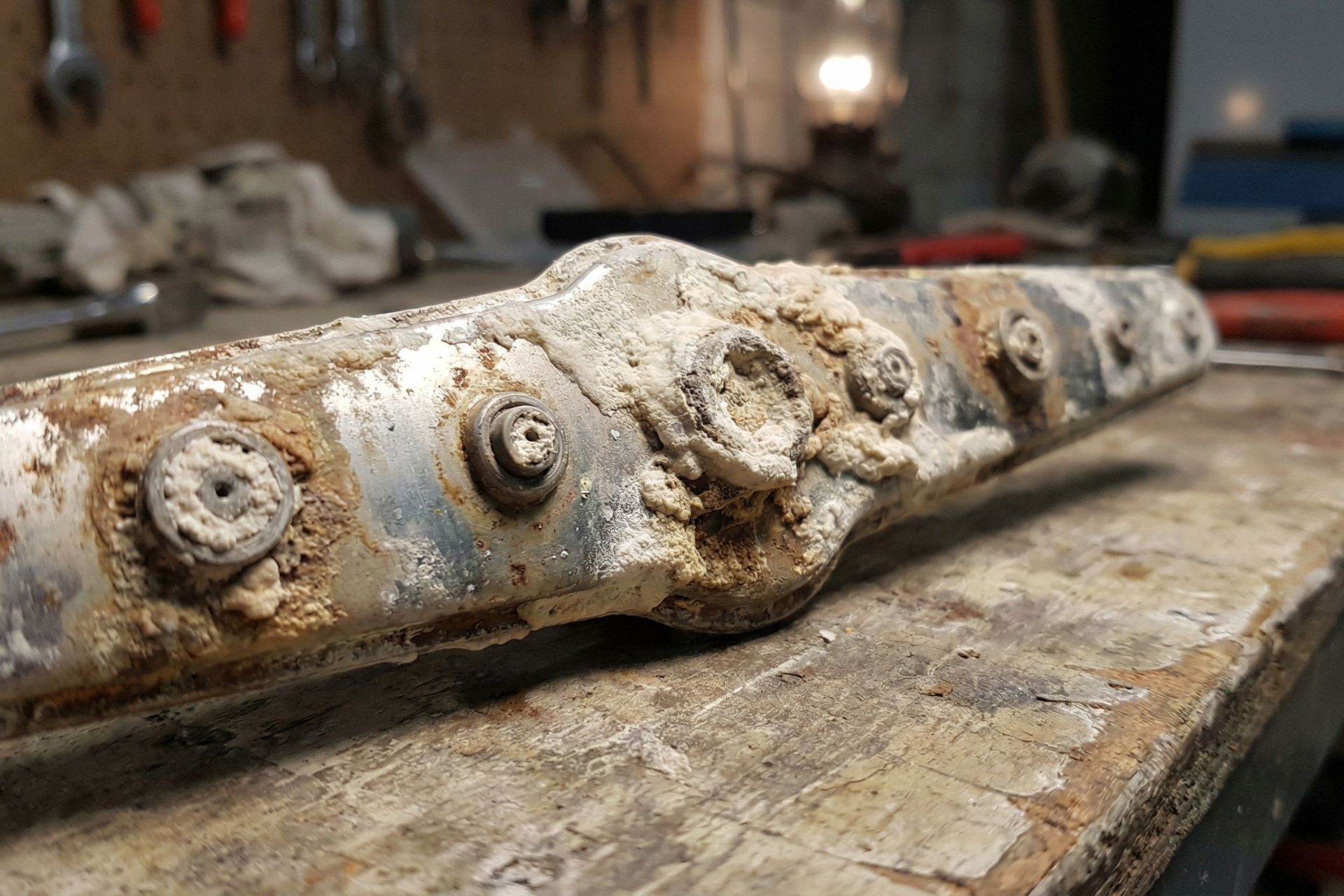 Close-up of severely corroded and scale-encrusted water pipe manifold or coupling showing extreme mineral buildup, rust, and deterioration on metal fittings laying on workbench in repair shop with tools in background.