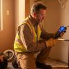 Plumber using digital leak detector on copper pipes during home inspection, wearing safety vest and gloves