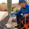 Technician using sewer line camera for residential drain inspection, viewing pipe video on tablet at outdoor cleanout