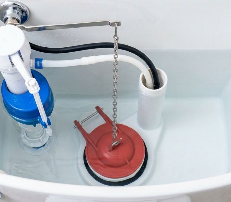 Toilet tank interior showing fill valve, overflow tube, and red flapper chain—DIY fix for running toilet plumbing repair.